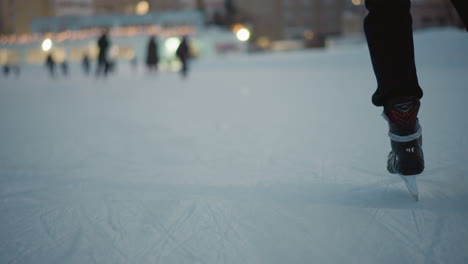 back view skater gliding gracefully on ice in winter evening wearing black skates with blurred distant people and glowing bokeh lights of buildings in background under soft fading daylight