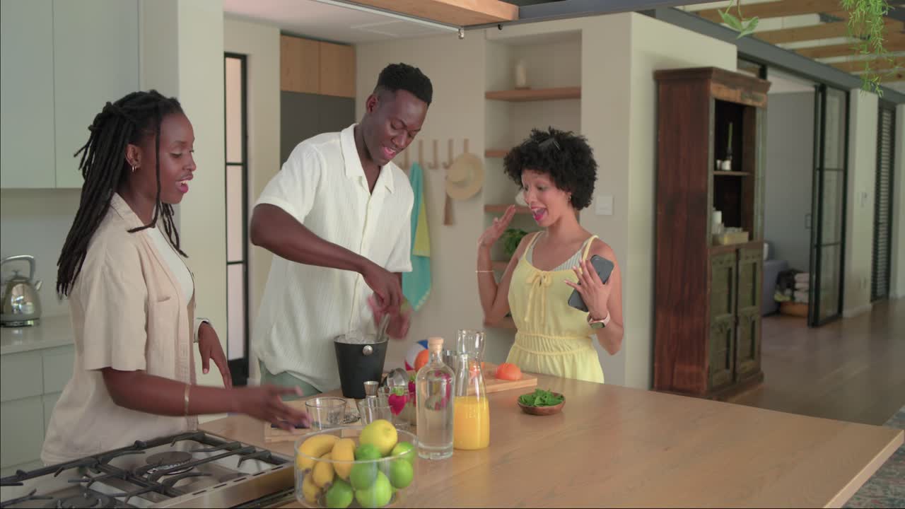 Amigos afro-americanos cortando laranjas, segurando o celular, interagindo e preparando bebidas na ilha da cozinha.