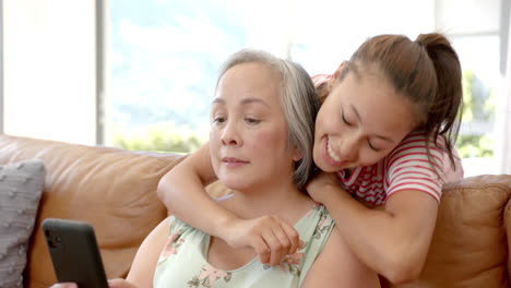 using smartphone, asian grandmother sitting on couch with granddaughter hugging her