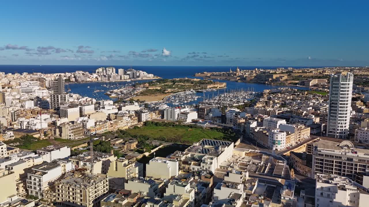 Aerial timelapse of Malta's urban skyline, showcasing Ta' Xbiex, Valletta, Sliema, Manoel Island, marina, and the Mediterranean Sea framed by a mix of modern architecture under a sunny winter sky