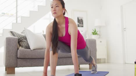 Asian-woman-on-mat-wearing-earphones,-exercising-with-smartphone-at-home