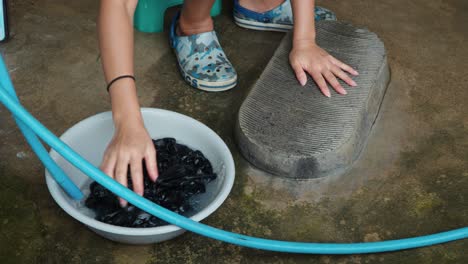 las manos de la mujer lavan enjuagar en un lavabo de plástico y retorcer la ropa en la tabla de lavado de losa vieja en el suelo de hormigón en una aldea pobre