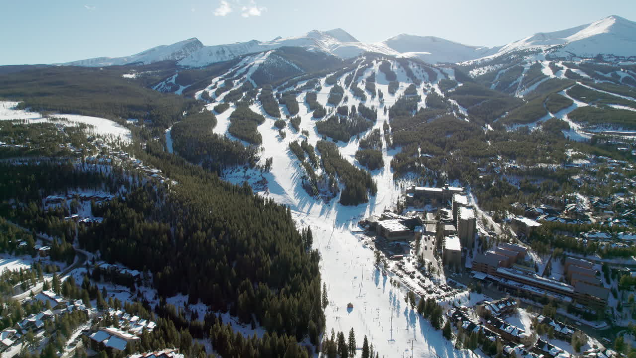 An aerial view approaching the popular Breckenridge ski resort in Colorado, with snow covered mountain peaks and high altitude trails full of winter skiers and snowboarders on a sunny winter day.