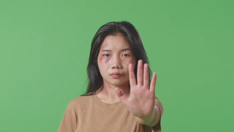 close up of young asian woman with bruise on face looks into camera showing hand sign to stop violence in the green screen background studio