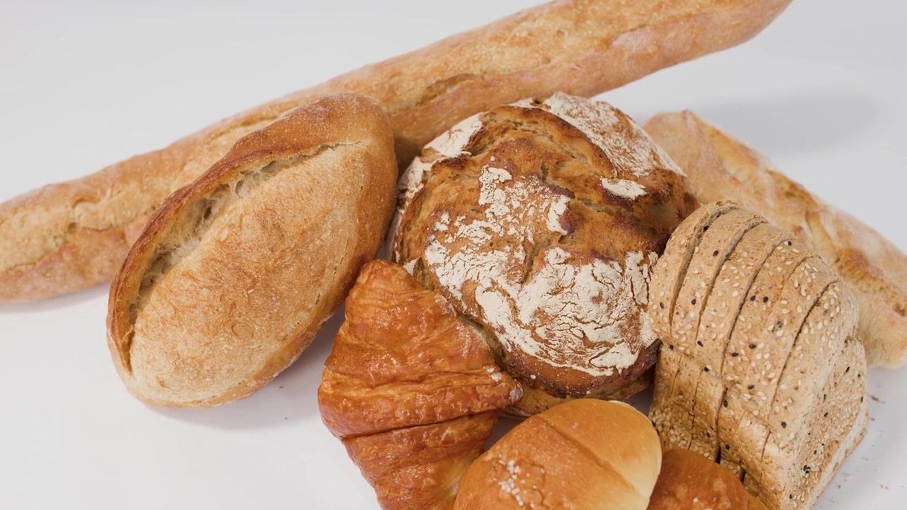 Slow camera zoom on variety of crusty baguettes, sourdough loaves, and croissants against white.