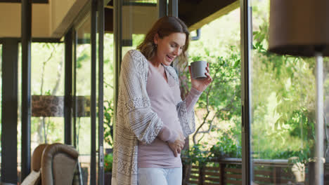 Glückliche-Kaukasische-Schwangere-Frau,-Die-Mit-Einer-Tasse-Tee-Am-Fenster-Steht-Und-Den-Bauch-Berührt