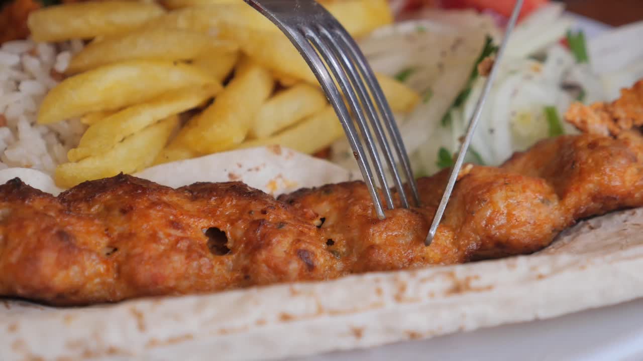 A close-up of a person cutting a kebab with fries, rice, and flatbread on a plate