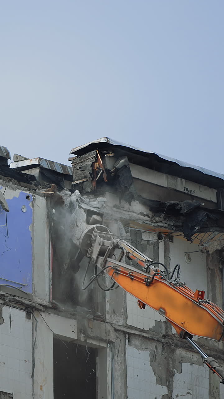 Excavator claw breaks the parts of ruined building. Dangerous deconstruction site with falling debris. Demolishing of the upper floors. Vertical video