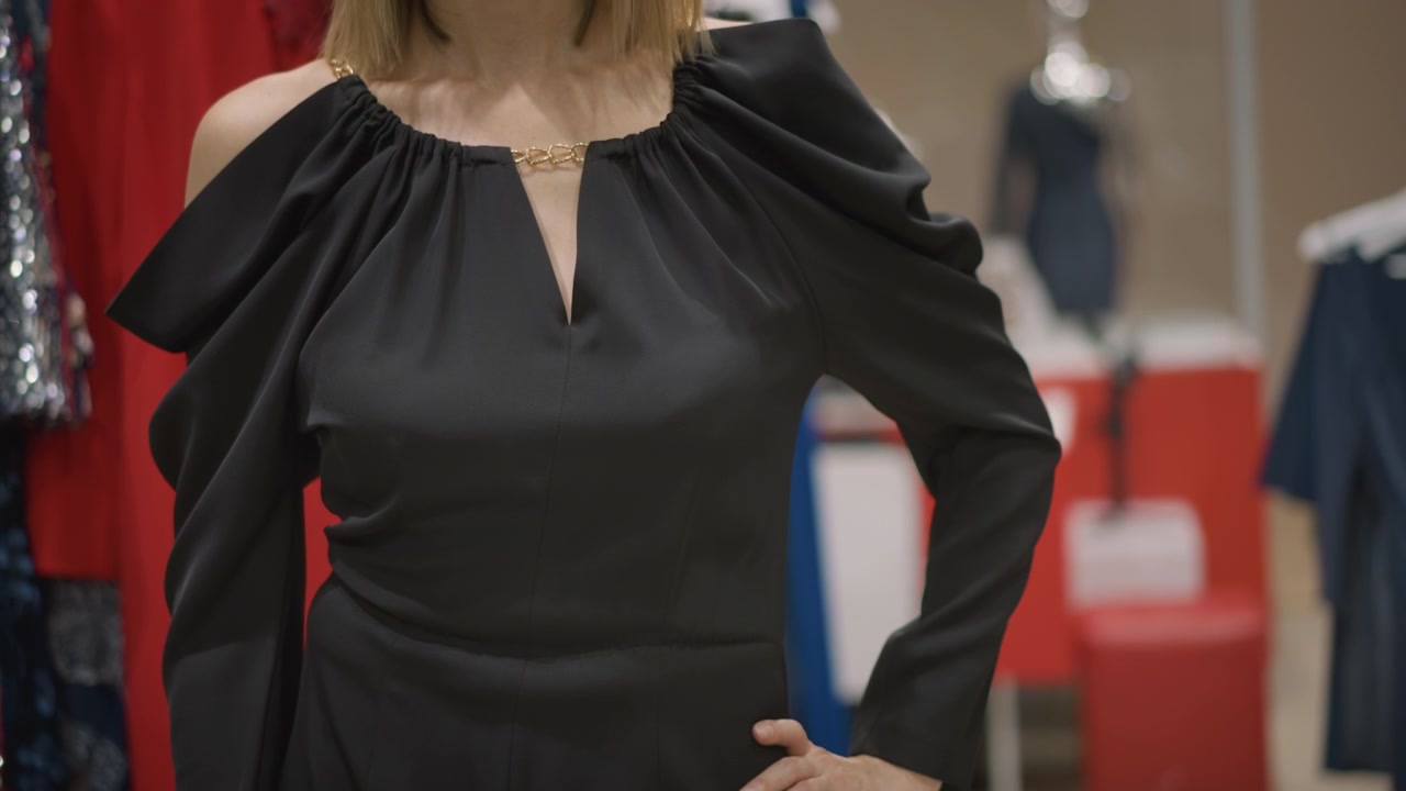 Primo piano di donna che indossa abiti neri in un ambiente interno, mostrando la parte inferiore del corpo e le mani. Il soggetto è immobile, con movimenti minimi, circondata da vari tessuti e capi d'abbigliamento in esposizione.