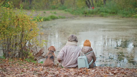 lady holding dog leash sits beside daughter on fallen leaves near peaceful lake surrounded by autumn foliage sharing tranquil moment together in nature enjoying stillness and soft light of forest edge