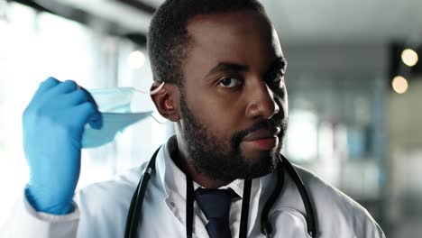 Close-up-of-African-American-physician-taking-off-medical-mask-and-goggles-and-looking-at-camera-in-clinic