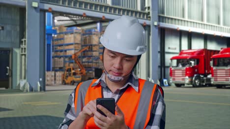 close up of asian male engineer with safety helmet using smartphone while standing , outside of logistics distributions warehouse