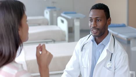 happy diverse male doctor and female patient sitting in hospital ward talking, slow motion