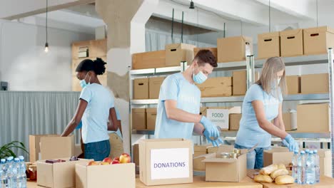 multiethnic group of volunteers in facial mask packing boxes with food and clothes in charity warehouse