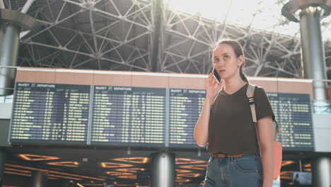 Parada-En-La-Pantalla-Con-Información-Sobre-La-Salida-Del-Avión-En-El-Aeropuerto,-Una-Joven-Con-Una-Mochila-Hablando-Por-Un-Teléfono-Móvil