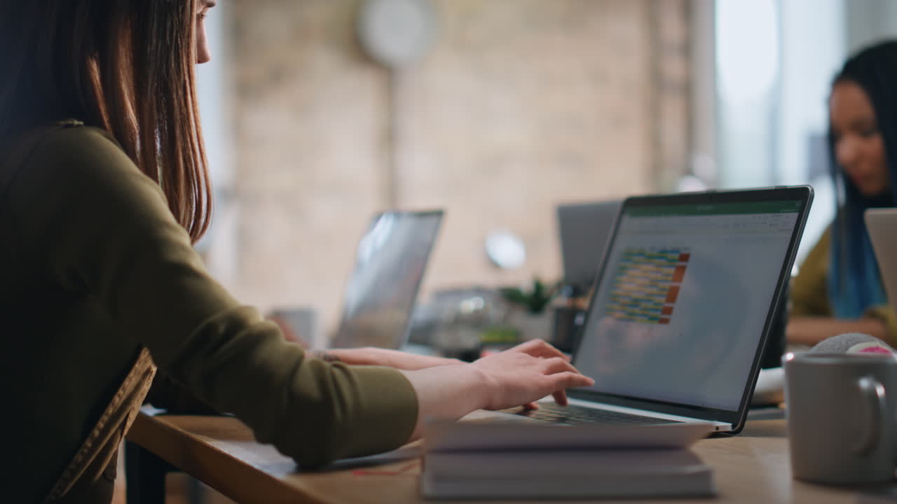 Ung kvinde skriver på bærbar computer i et coworking space, tæt på. Leder skriver rapport.