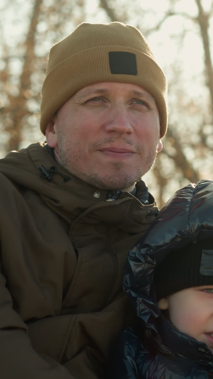 primer plano de un padre y un hijo sentados juntos en un banco, con el padre mirando enfocado, los suaves rayos del sol se reflejan suavemente detrás de él, atmósfera, con un fondo borroso de árboles