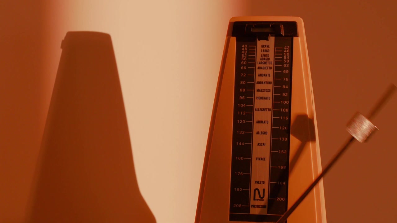 Nahaufnahme eines Vintage-Metronoms mit silbernem Pendel, das im langsamen Rhythmus mit achtzig Schlägen pro Sekunde in einem schummrigen Raum mit orangefarbenem Hintergrund schlägt. Das Metronom wirft einen Schatten an die Wand.