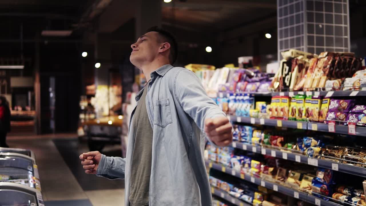 슈퍼마켓에서: 행복한 세련된 남자가 상품을 가진 선반 앞에 큰 쇼핑몰에서 자유롭게 춤을 춘다.