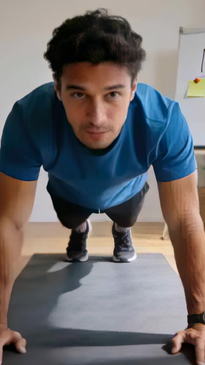 Video verticale: Uomo che inizia a fare flessioni su tappetino grigio per fitness a casa, con maglietta sportiva e lavagna bianca.