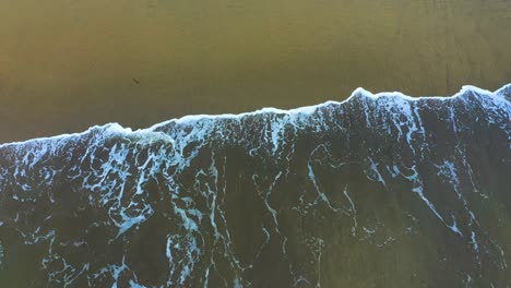 a drone captures a dramatic view of the ocean's energy as waves crash onto the sandy shore, creating frothy white foam