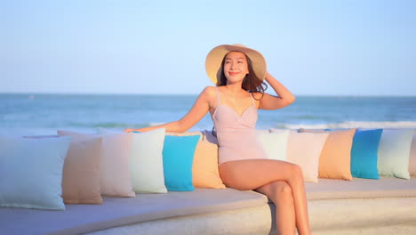 Sexy-Asian-model-wearing-purple-swimming-suit-and-straw-floppy-hat-sits-on-the-beach-stone-bench-seaside-smiling-relaxing-on-outdoor-patio-furniture-on-Thailand-vacation-at-resort-terrace