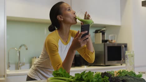 mixed race woman drinking healthy drink and using smartphone in kitchen