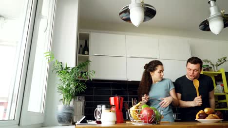 una joven pareja alegre se divierten bailando y cantando mientras preparan el desayuno en la cocina de casa.