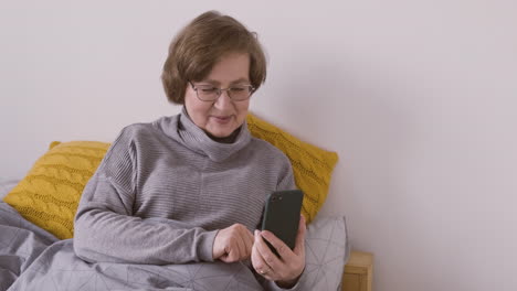 senior woman with eyeglasses sitting on bed making a video call on a smartphone