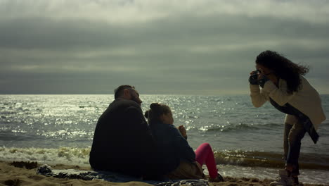 Familia-Feliz-Tomando-Fotografías-En-La-Playa-Del-Océano.-Gente-Sentada-Posando-Cámara-Junto-Al-Mar.