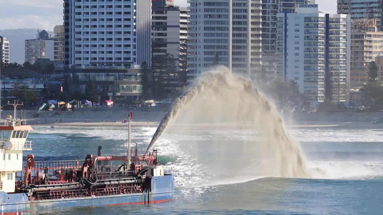 A dredger pumps sand onto the beach, set against Gold Coast's urban skyline. Bright daylight enhances the dynamic scene