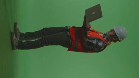 full body side view of asian male engineer with safety helmet working on a laptop while standing in the green screen background studio, during sunset or sunrise time