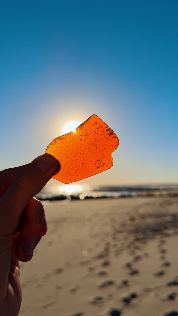 Una mano che tiene un pezzo di vetro arancione su una spiaggia durante il tramonto. Una persona solleva un pezzo di vetro arancione brillante sullo sfondo di un tramonto sulla spiaggia.