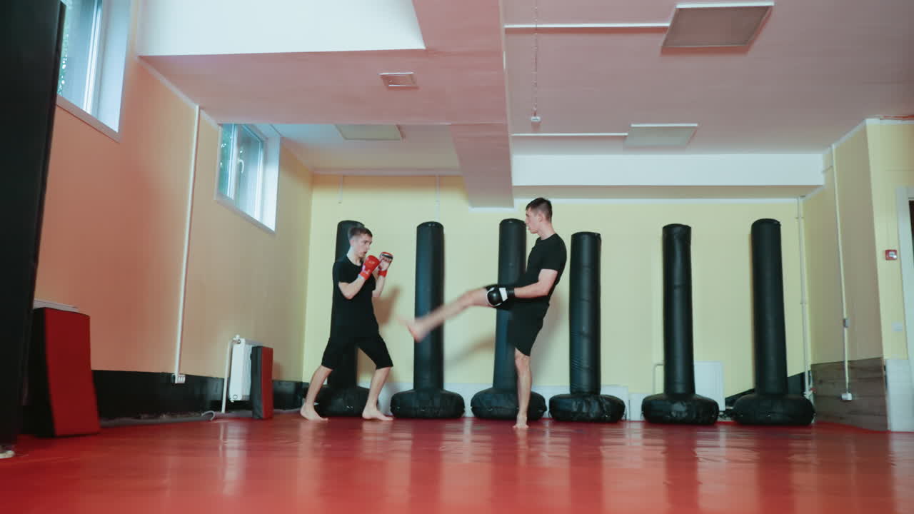 Two wrestlers spar barefoot on red mat in gym, one throwing punch while opponent blocks near wall lined with black punching bags, showcasing strength, focus, energy, martial arts training intensity