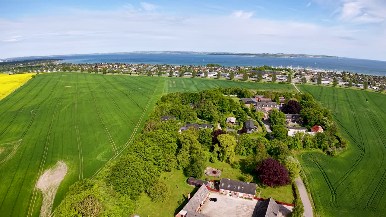 Ilmakuvaus droneilla rehevistä vihreistä viljelysmaista, joilla on maatiloja ja pieni rannikkokaupunki lähellä merta Tanskassa