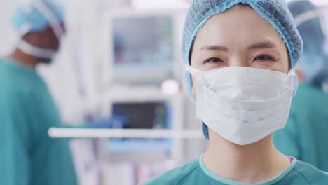 video portrait of smiling asian female surgeon wearing face mask in operating theatre, copy space