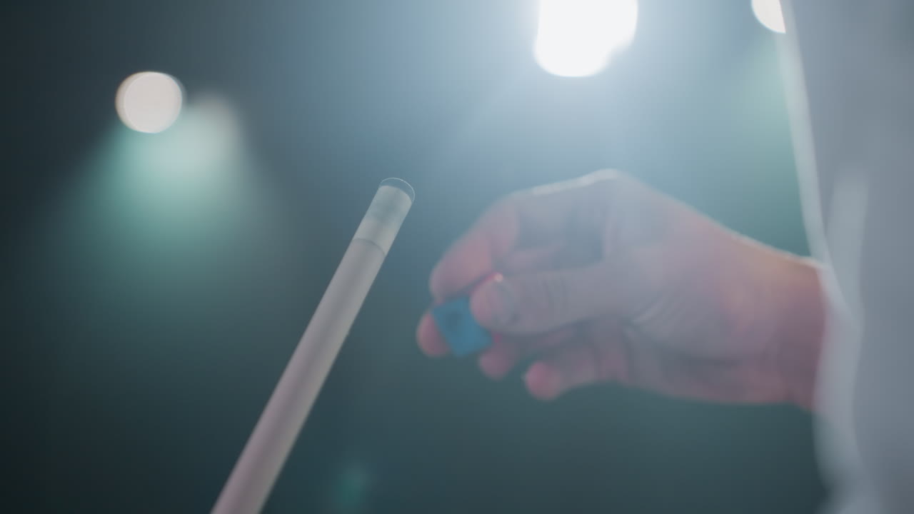 손가락이 조심스럽게 빨간색과 파란색 도자기를 빌리어드 게임 전에 큐 스틱에 적용하는 클로즈업. 집중된 손잡이와 흐릿한 배경은 정밀성, 집중력을 강조합니다.