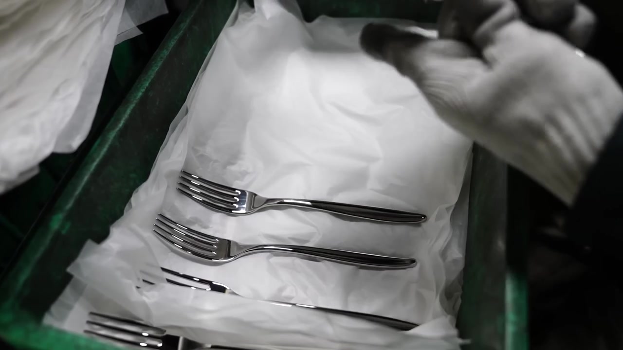 Worker in white gloves laying finished forks onto tissue paper inside green plastic crate for transport
