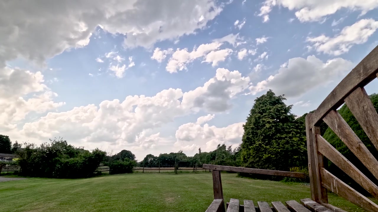 Wolken ziehen über den blauen Himmel über einem grünen Garten mit Bäumen und einer Holzbank. Zeitraffer.