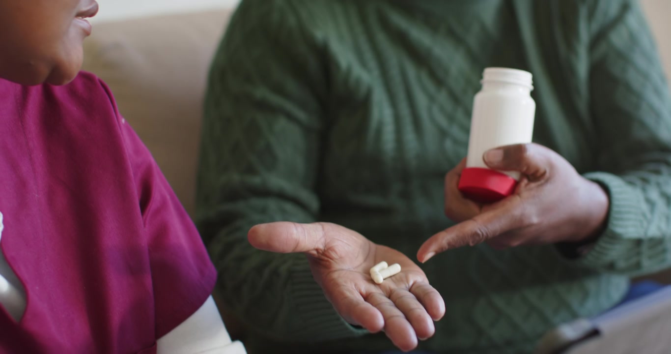 During meds review man pouring pills from bottle into caregiver hand while checking tablet on sofa