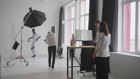 Art-director-checking-the-photos-on-a-monitor.-Professional-crew-team-together-in-the-studio.-Behind-the-scene-of-photo-shooting-Professional-football-player-and-production-set-up-in-the-big-studio.