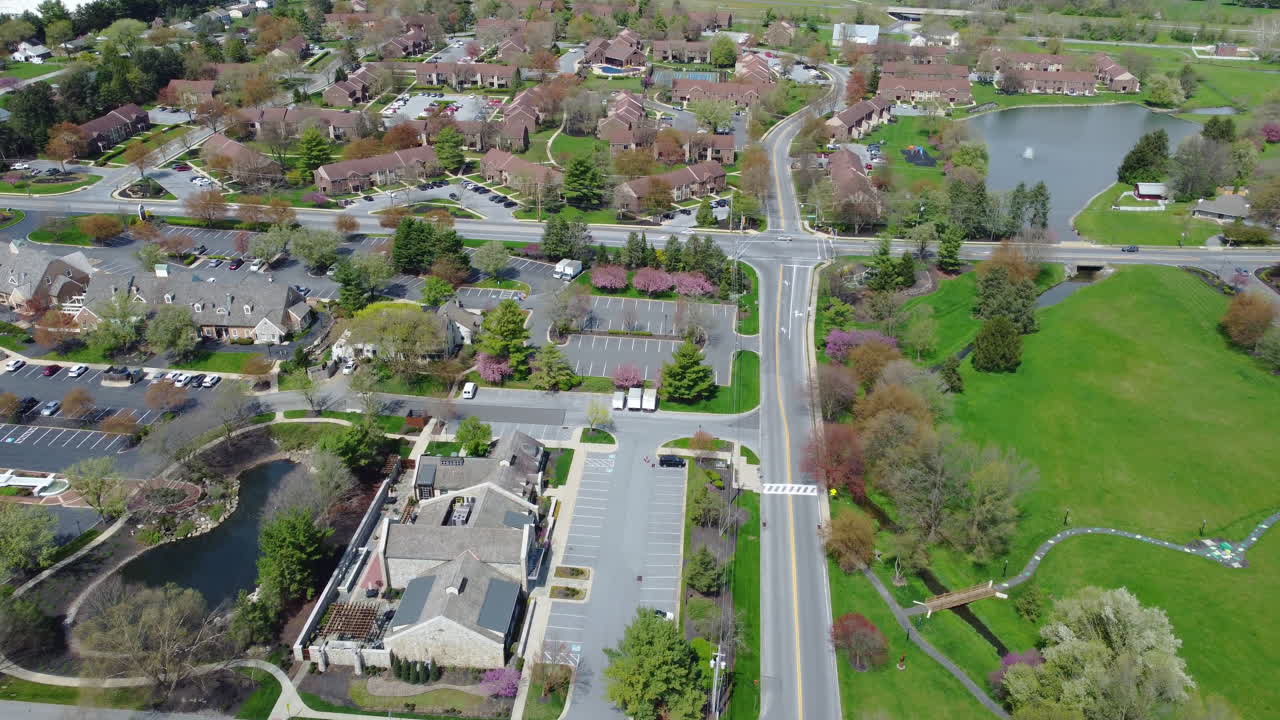 A drone glides along a road transitioning from a quiet commercial office area into a landscaped residential neighborhood in Lancaster, PA. Spring foliage and community design blend urban and suburban.