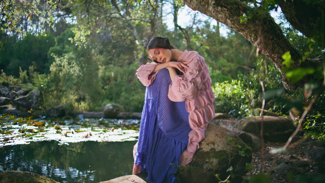 마법의 여자가 자연의 평온을 즐기며 숲의 연못 근처에 앉아 있습니다. 로맨틱한 소녀 포즈