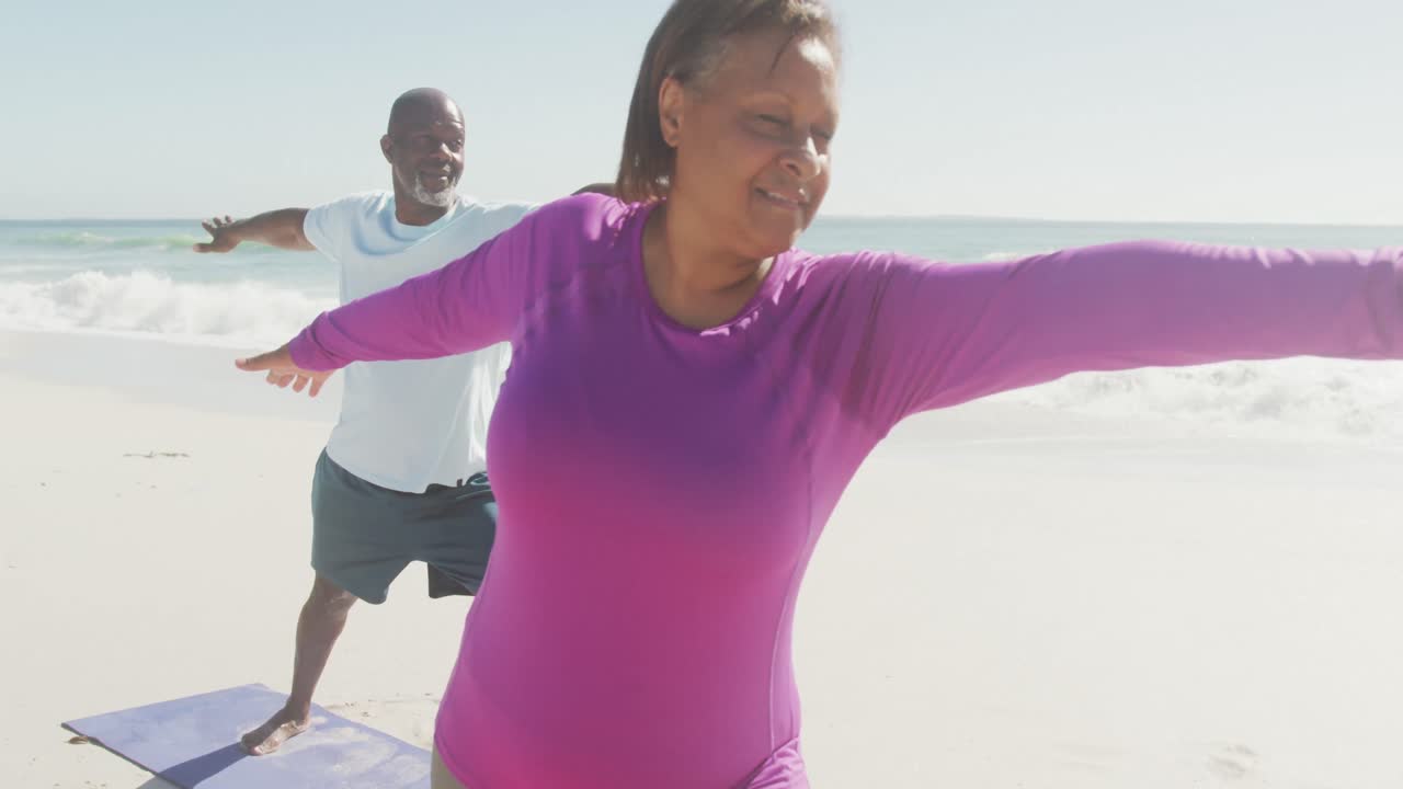 una pareja afroamericana mayor sonriendo y practicando yoga en esteras en una playa soleada
