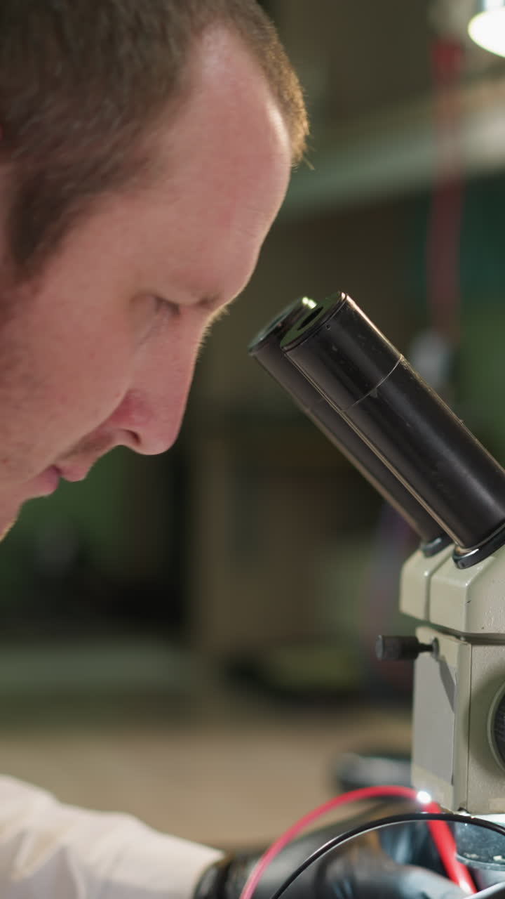 Middle view of a technician in a white lab coat and black gloves carefully testing a circuit under a microscope using a voltmeter in his laboratory, with the desk lamp on