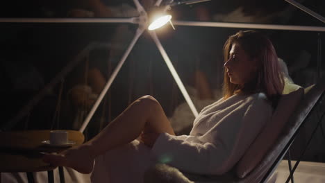mujer elegante vestida con túnica de baño pone la pierna sobre la pierna sentada en la mesa. dama seria descansa junto a la ventana panorámica con una taza de bebida caliente disfrutando de una agradable noche