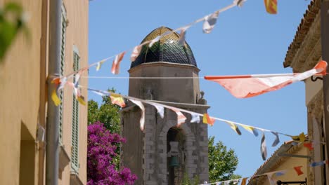 lleno de banderitas de colores ondeando en el viento, atado en una cuerda colgando entre dos paredes de un callejón con una iglesia y su campana en el fondo, clima soleado, cielo azul
