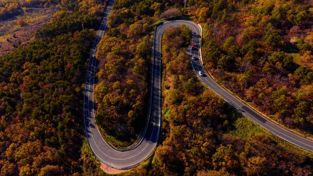 Letecký záběr shora na klikatou silnici, která se vine hustým lesem s živým podzimním listím v oranžových a zlatých tónech