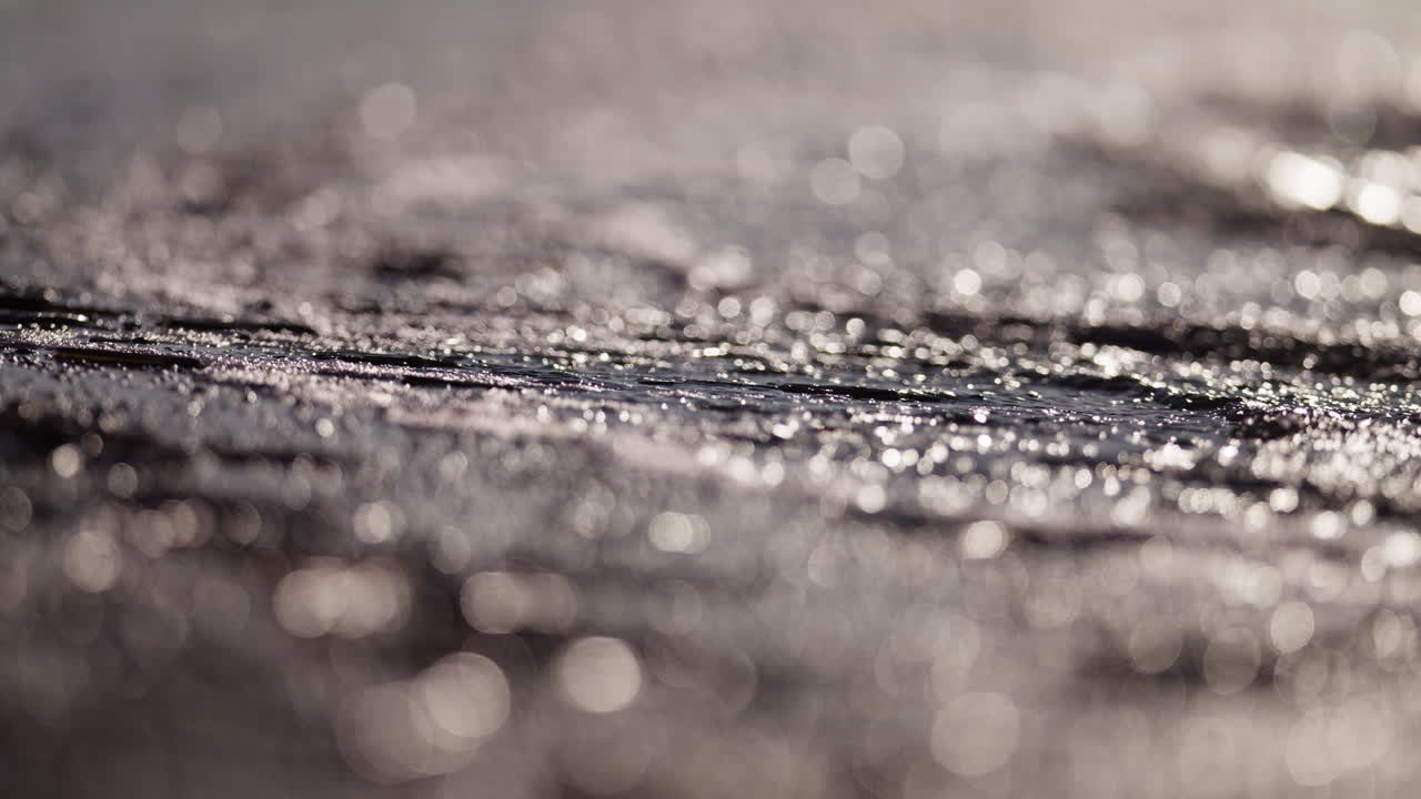 Sunset light shimmers across calm ocean surface forming warm reflective patterns near shore, telephoto abstract backdrop, slow motion