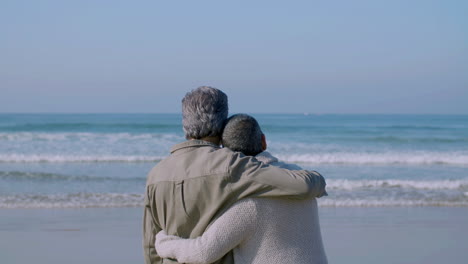 Senior-couple-hugging-on-beach-and-looking-at-ocean